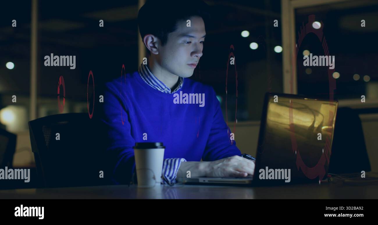 Homme chinois travaillant dans le pull bleu tapant sur ordinateur portable tard dans la nuit au bureau, avec tasse à café Banque D'Images