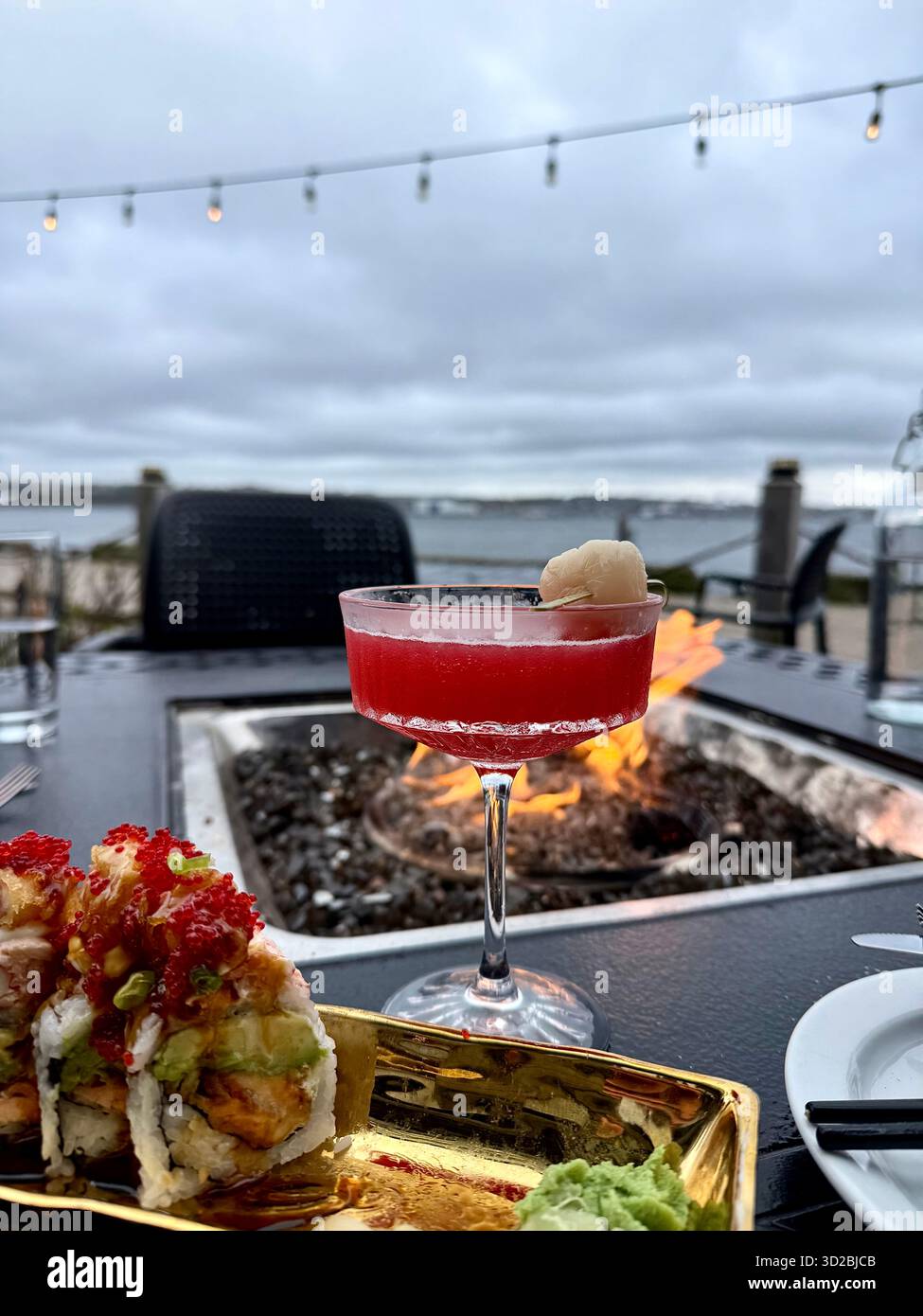 Restaurant gastronomique en plein air avec une assiette de rouleaux de sushi et un cocktail rouge garni de litchi, sur une table à côté d'un foyer confortable. Le ca - Image de stock capturée avec un smartphone