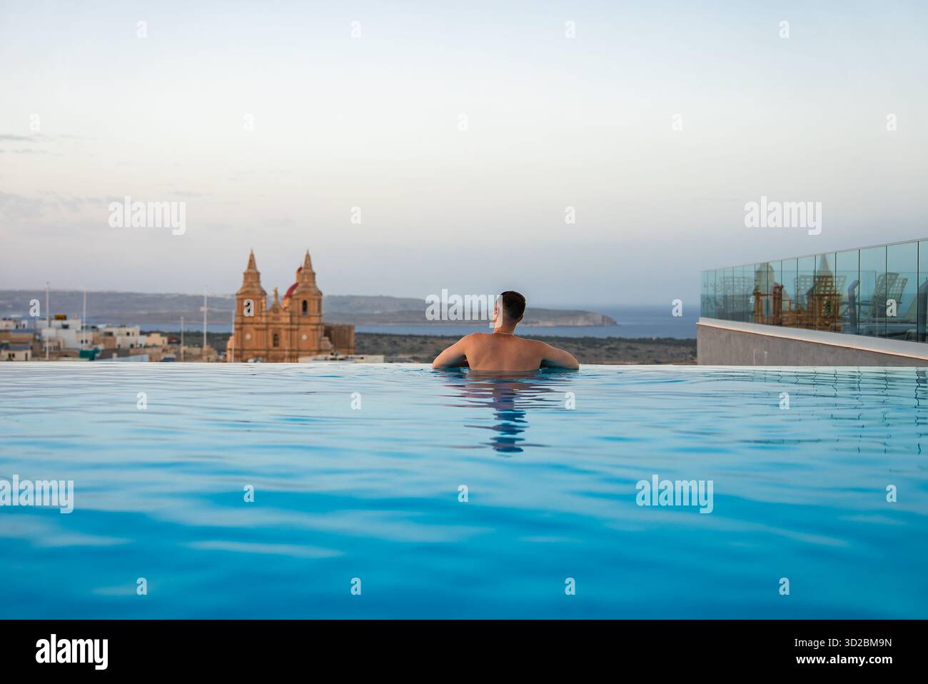 Une personne se repose près d'une piscine à débordement sur le toit à Mellieha, Malte au crépuscule, avec le Sanctuaire de notre-Dame de Mellieha et Comino et Gozo visible au-delà de CA Banque D'Images