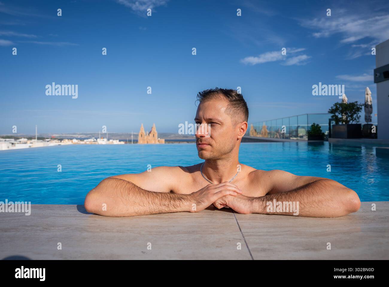 Un homme repose dans une piscine à débordement sur un toit à Mellieha, Malte. Il fait face à l'horizon, avec l'église paroissiale Mellieha, Comino et Gozo au-delà dans Crisp w Banque D'Images