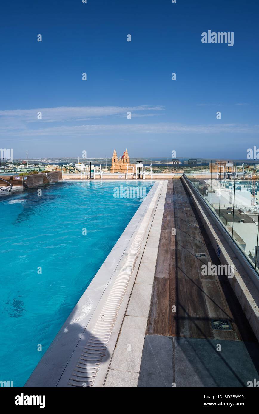 Une piscine à débordement sur le toit longe une balustrade en verre à Mellieha, Malte. L'église paroissiale Mellieha s'élève au-delà, avec la Méditerranée, Comino et Gozo ier Banque D'Images