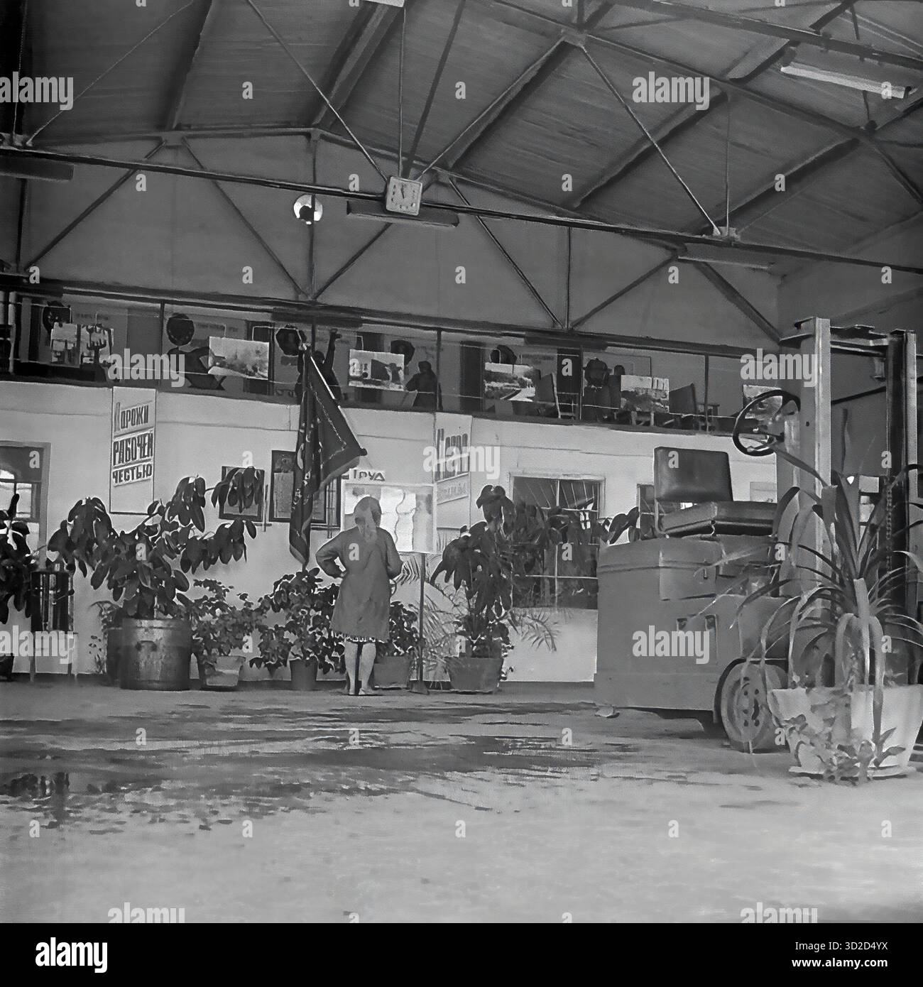 Archive photo en noir et blanc des années 1970 à Sloviansk, RSS d'Ukraine. Il montre un hall spacieux ou un dépôt à l'armature-Insulator Plant (AIZ). Une femme se tient au centre. Un chariot élévateur est stationné sur la droite. Le fond a une mezzanine (deuxième niveau) servant de « coin rouge » ou de bureau, décoré d'affiches de propagande soviétique et d'un drapeau. La salle est fortement décorée avec de grandes plantes en pot, typiques de l'embellissement de l'usine soviétique. Banque D'Images