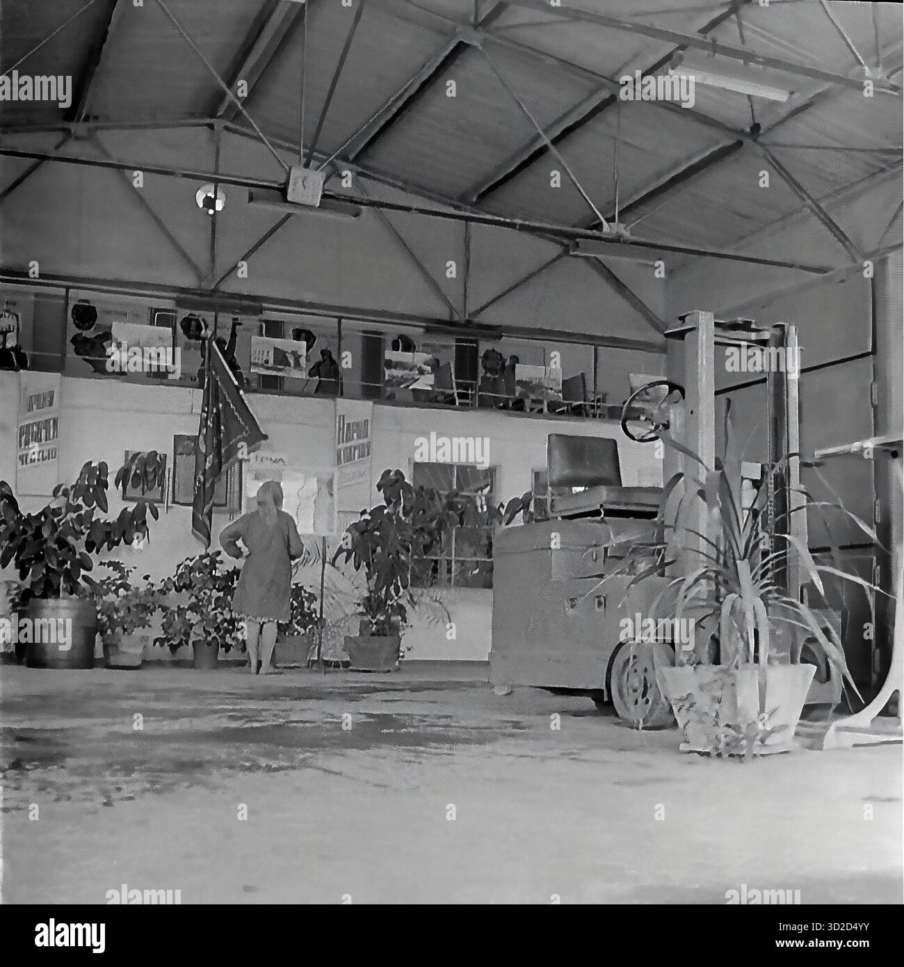 Archive photo en noir et blanc des années 1970 à Sloviansk, RSS d'Ukraine. Il montre un hall spacieux ou un dépôt à l'armature-Insulator Plant (AIZ). Une femme se tient au centre. Un chariot élévateur est stationné sur la droite. Le fond a une mezzanine (deuxième niveau) servant de « coin rouge » ou de bureau, décoré d'affiches de propagande soviétique et d'un drapeau. La salle est fortement décorée avec de grandes plantes en pot, typiques de l'embellissement de l'usine soviétique. Banque D'Images