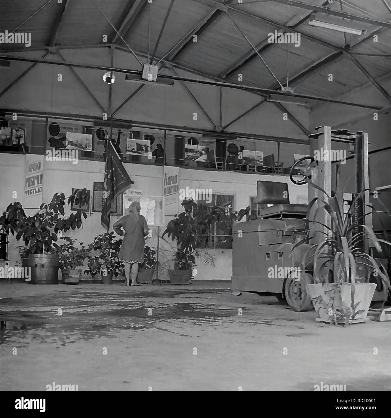 Archive photo en noir et blanc des années 1970 à Sloviansk, RSS d'Ukraine. Il montre un hall spacieux ou un dépôt à l'armature-Insulator Plant (AIZ). Une femme se tient au centre. Un chariot élévateur est stationné sur la droite. Le fond a une mezzanine (deuxième niveau) servant de « coin rouge » ou de bureau, décoré d'affiches de propagande soviétique et d'un drapeau. La salle est fortement décorée avec de grandes plantes en pot, typiques de l'embellissement de l'usine soviétique. Banque D'Images