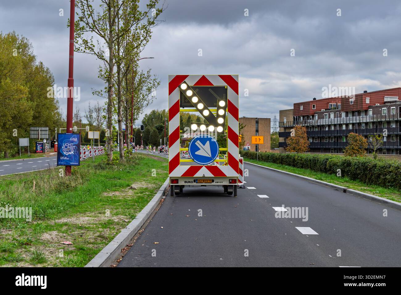 Un panneau fléché mobile sur une remorque jaune dirigeant la circulation à droite, garé sur un bord de route avec une bordure verte et des bâtiments modernes en arrière-plan. Utre Banque D'Images