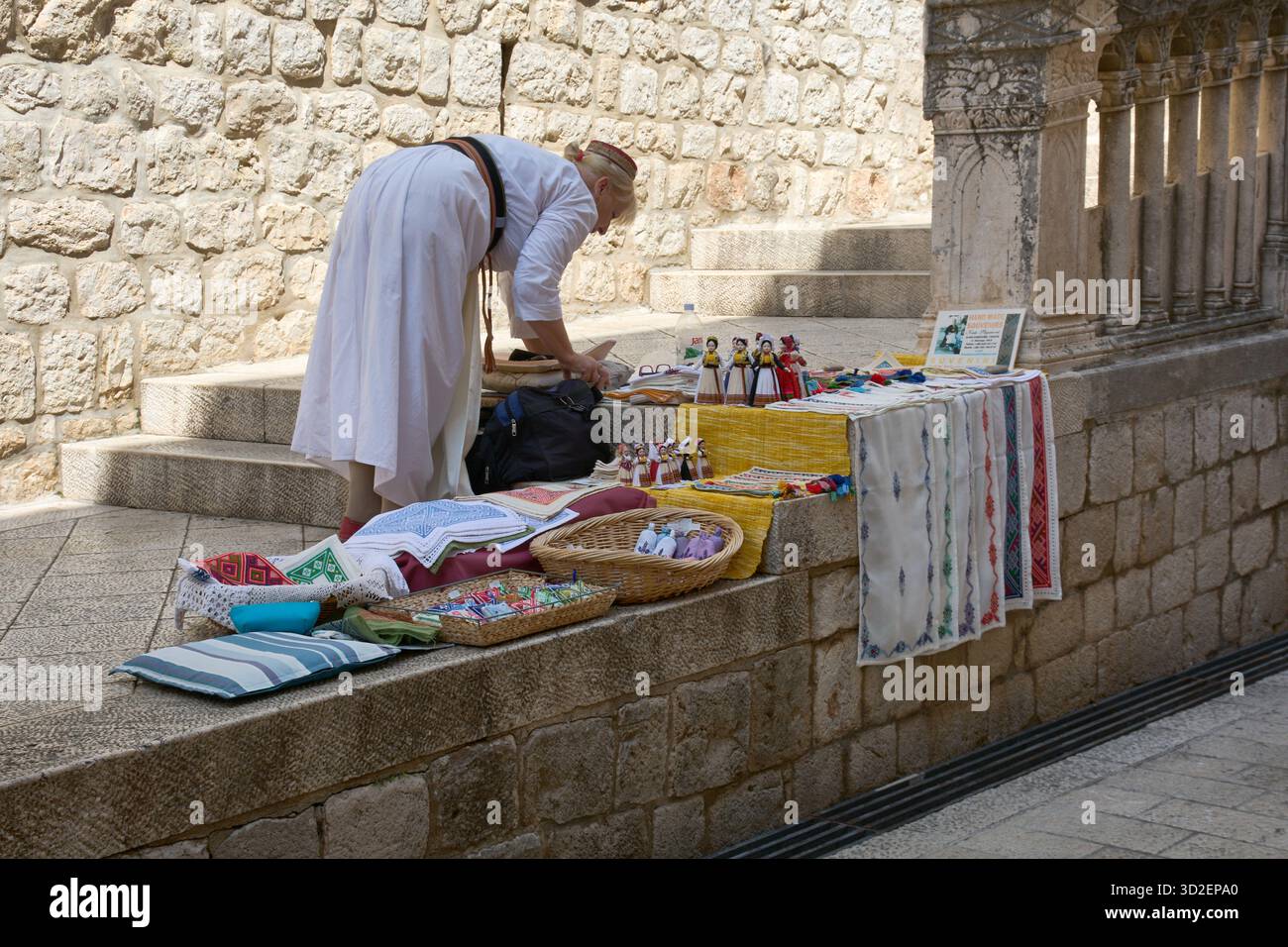 Dubrovnik. Croatie - 1er novembre 2025 : une vendeuse de rue en robe blanche traditionnelle met en place ses marchandises, y compris des textiles et des poupées, contre un salut Banque D'Images