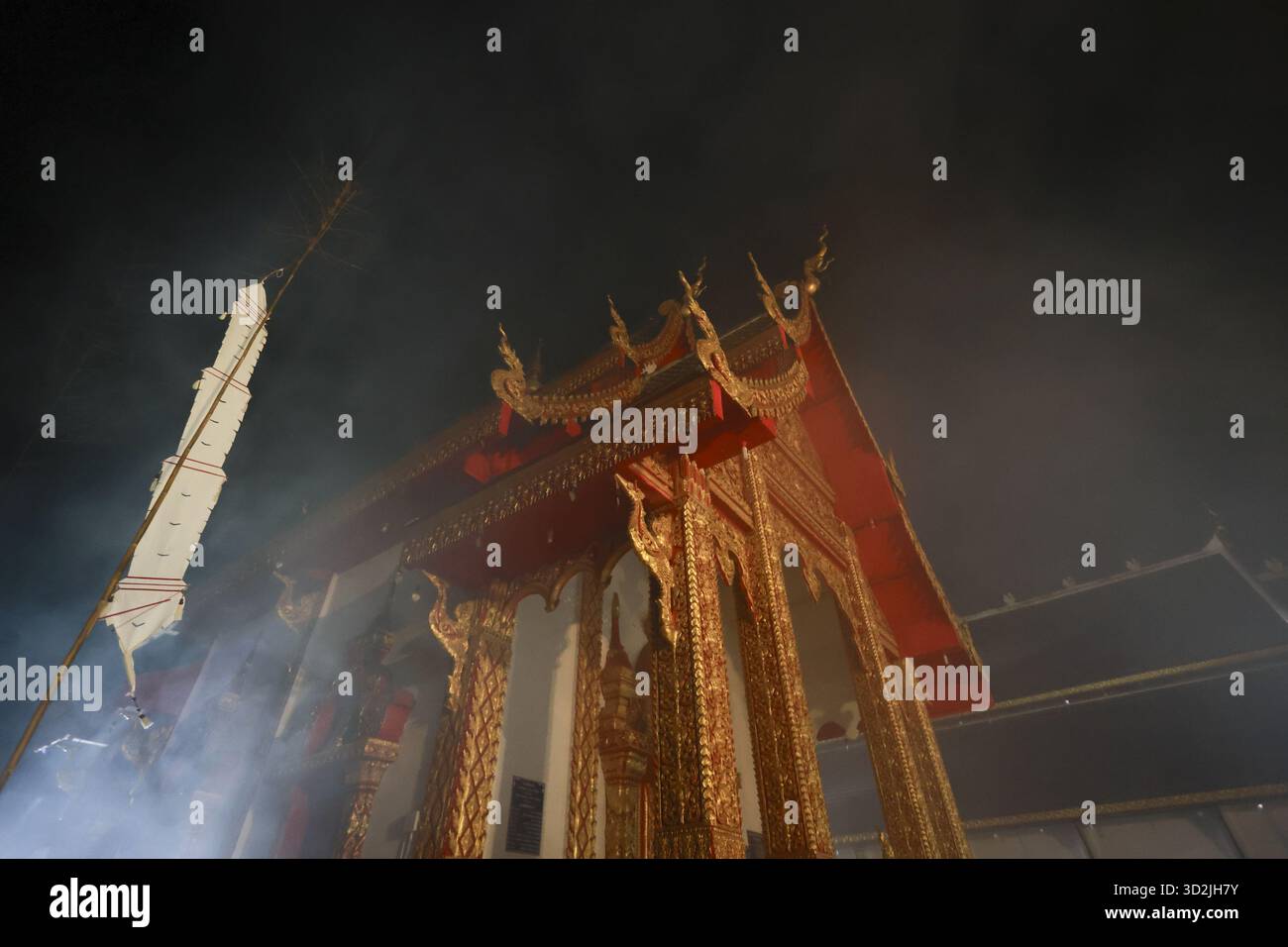 Temple thaïlandais doré sacré la nuit, enveloppé de fumée mystérieuse. Des rayons de lumière dramatiques illuminent l'architecture bouddhiste traditionnelle, créant du spirituel Banque D'Images