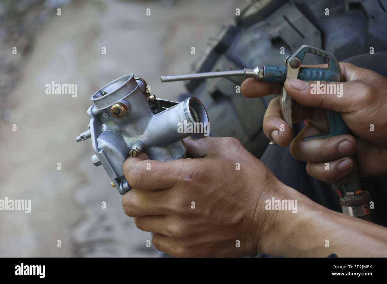 Mécanicien concentré avec la main sale effectue l'entretien de la moto, le nettoyage du carburateur argenté avec pistolet à air comprimé lors d'un service de réparation de moteur i Banque D'Images