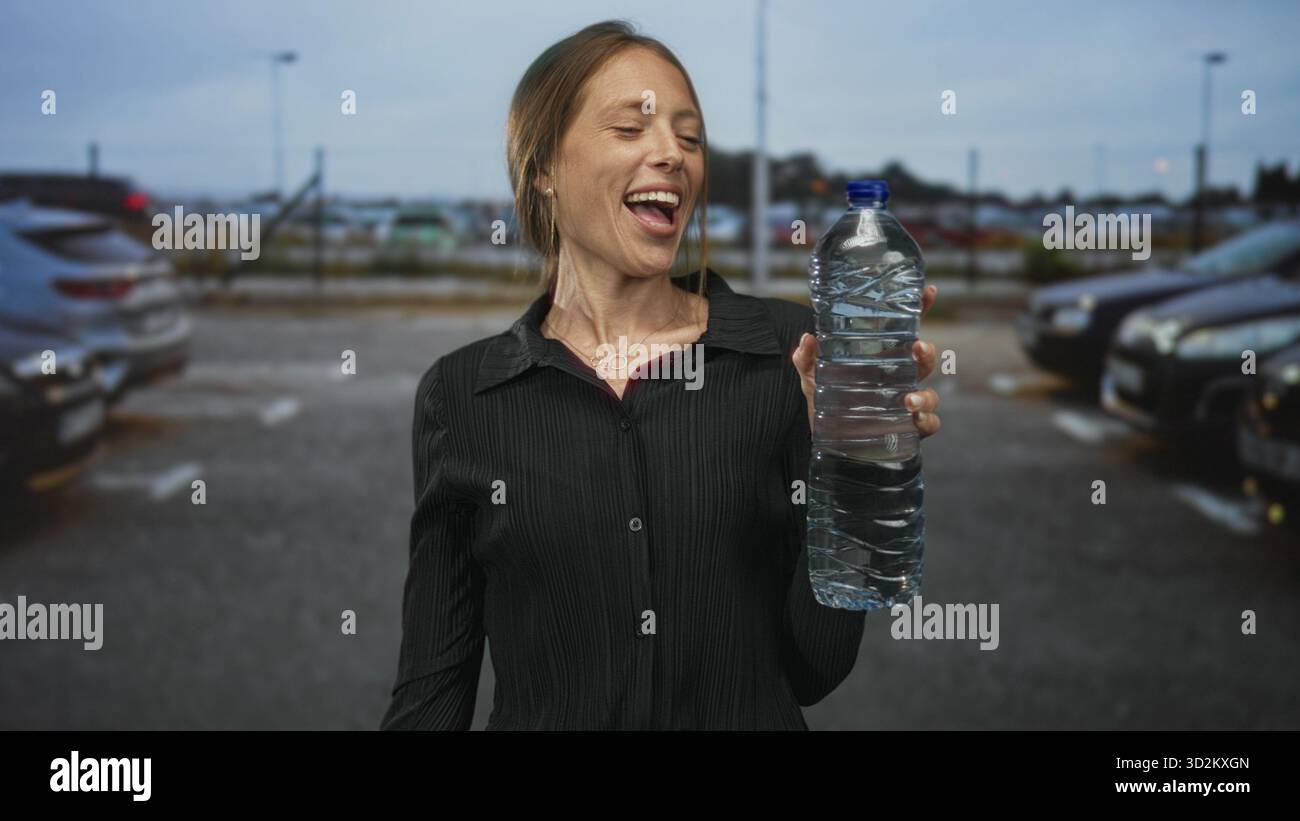 Femme tenant une grande bouteille d'eau et souriant tout en la regardant dans un parking de rue avec voitures et lampadaires visibles ; joie d'hydratation. Banque D'Images