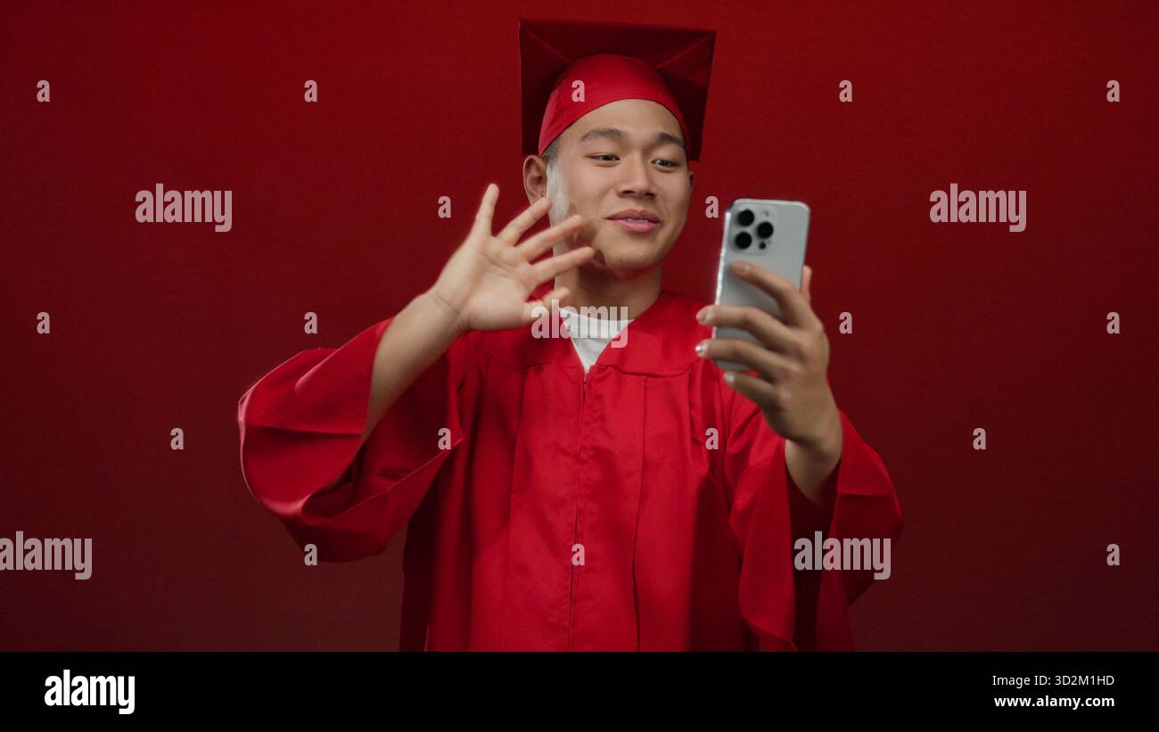 Jeune homme chinois étudiant en robe de graduation rouge faisant un appel vidéo sur smartphone au-dessus d'un mur rouge isolé célébrant la réussite scolaire avec une joyf Banque D'Images