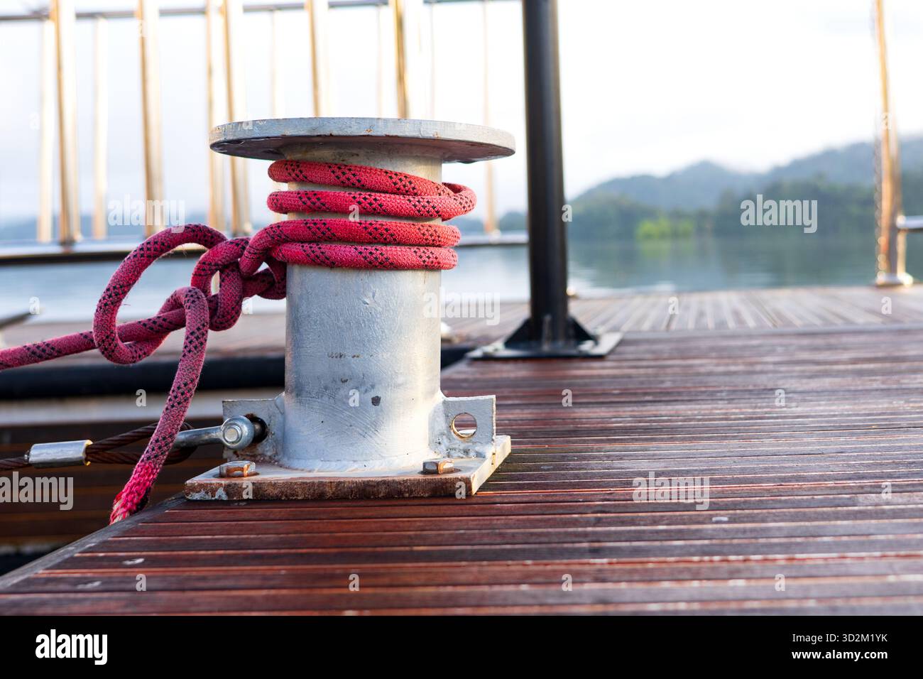 Borne en métal sur quai en bois avec corde rouge solidement enveloppée, attachant un bateau dans un cadre rustique Marina avec fond doux Banque D'Images