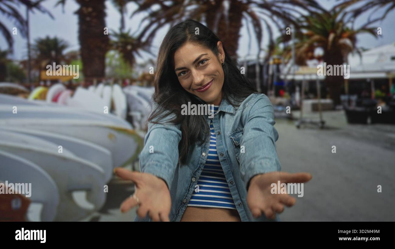 Femme avec les mains croisées sur la poitrine portant une veste en denim et un crop top rayé sur la rue avec des planches de surf et des palmiers ; connexion de gratitude. Banque D'Images