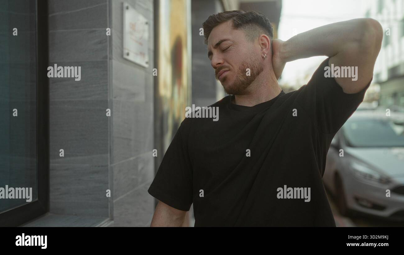 Jeune homme avec la barbe à l'extérieur dans la ville tenant le cou dans l'inconfort contre le fond urbain sous la lumière du soleil, portant chemise noire décontractée dans la rue pendant Banque D'Images