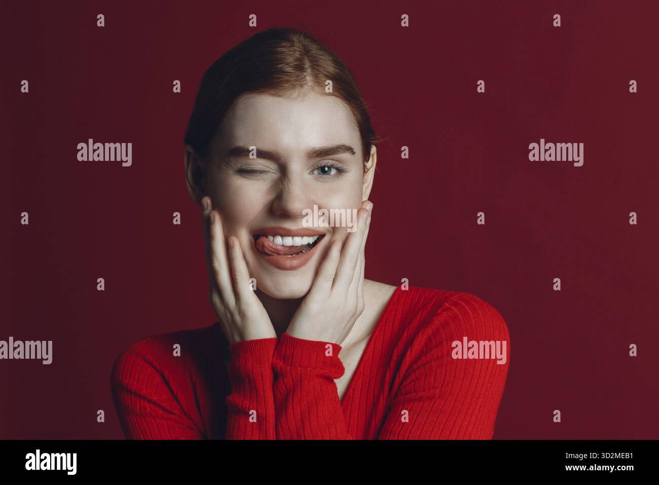 Portrait de jeune femme drôle de gingembre clin d'œil sur fond rouge avec les mains au visage et les expressions faciales Banque D'Images