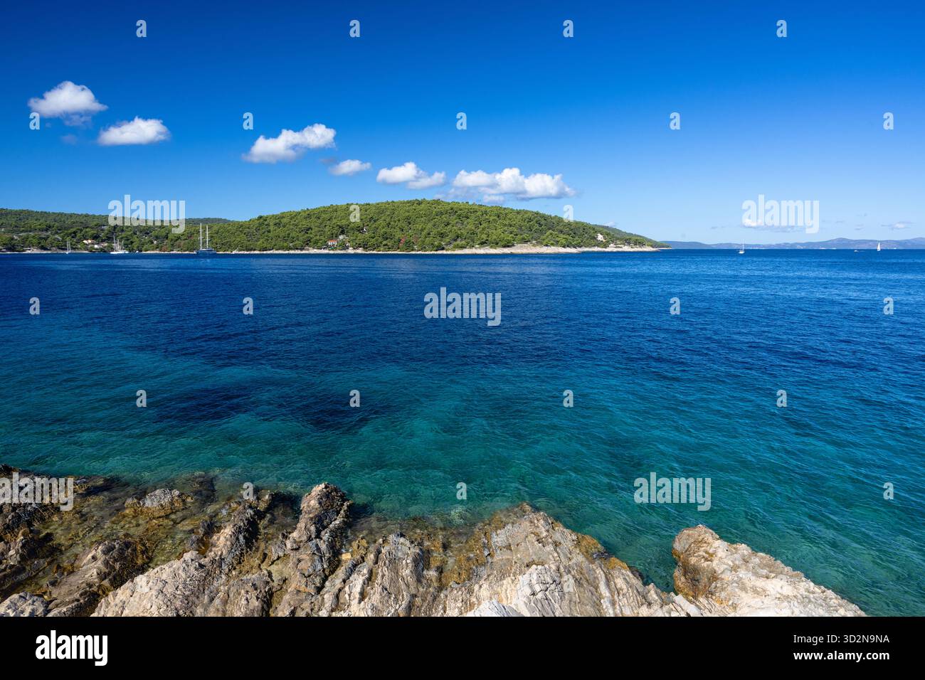 Un paysage marin tranquille avec un voilier solitaire glissant sur l'eau bleu profond, un premier plan rocheux, et une île verdoyante au loin sous un SK clair et lumineux Banque D'Images