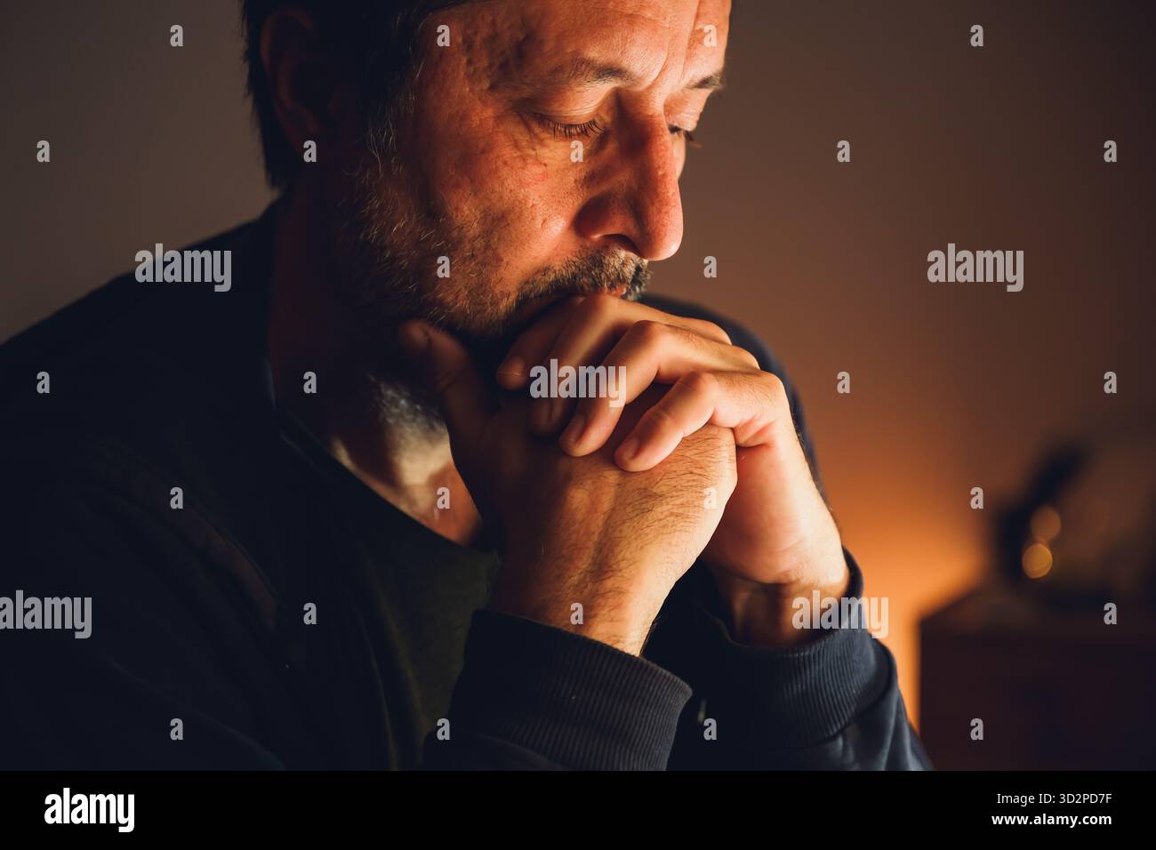 Homme inquiet assis seul dans une pièce sombre avec une lumière basse chaude, une expression sérieuse et une tension émotionnelle. Mise au point sélective. Banque D'Images