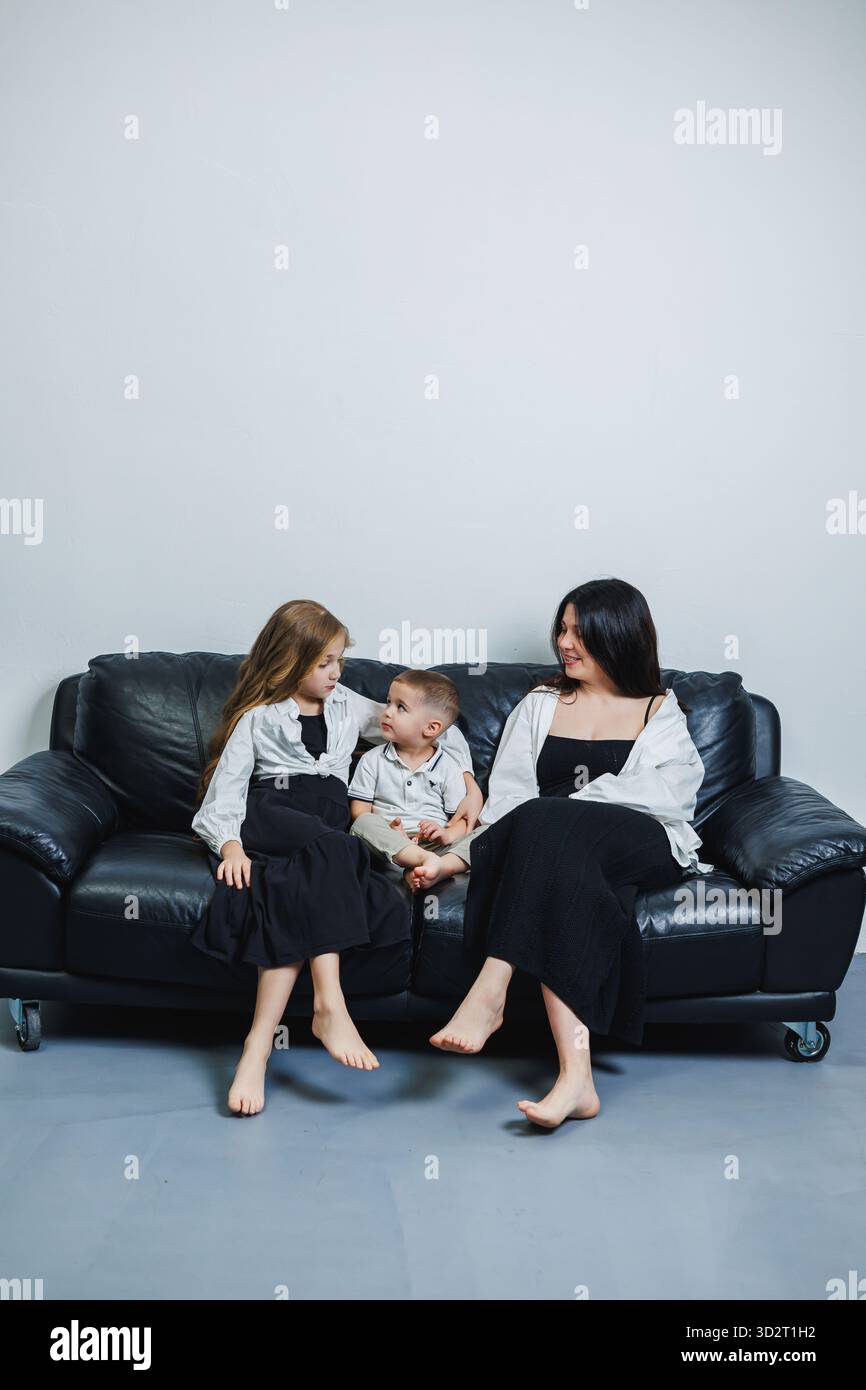 Une mère est assise sur un canapé noir avec ses deux enfants, riant et discutant. Les enfants sont habillés dans des tenues assorties, créant un guichet automatique chaleureux et joyeux Banque D'Images
