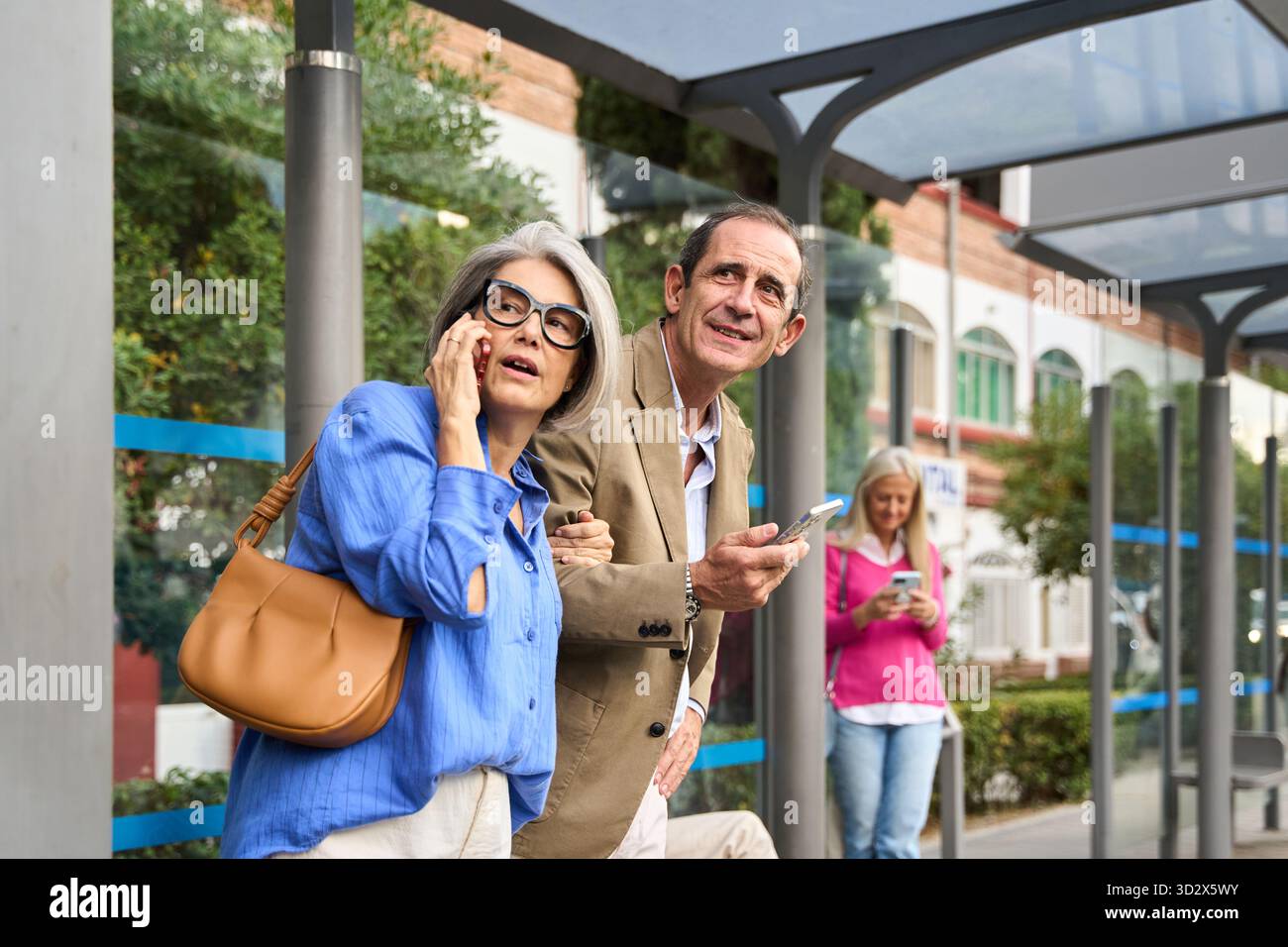 Couple senior attendant à un arrêt de bus de la ville, une femme parlant sur son téléphone, un homme vérifiant son smartphone Banque D'Images