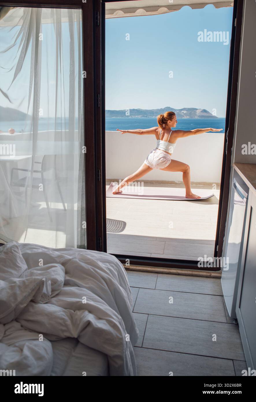 Femme d'âge moyen faisant le yoga du matin guerrier exercice d'équilibre de posture sur la vue mer Terace chambre d'hôtel, faisant Virabhadrasana pose qui rend votre corps mor Banque D'Images