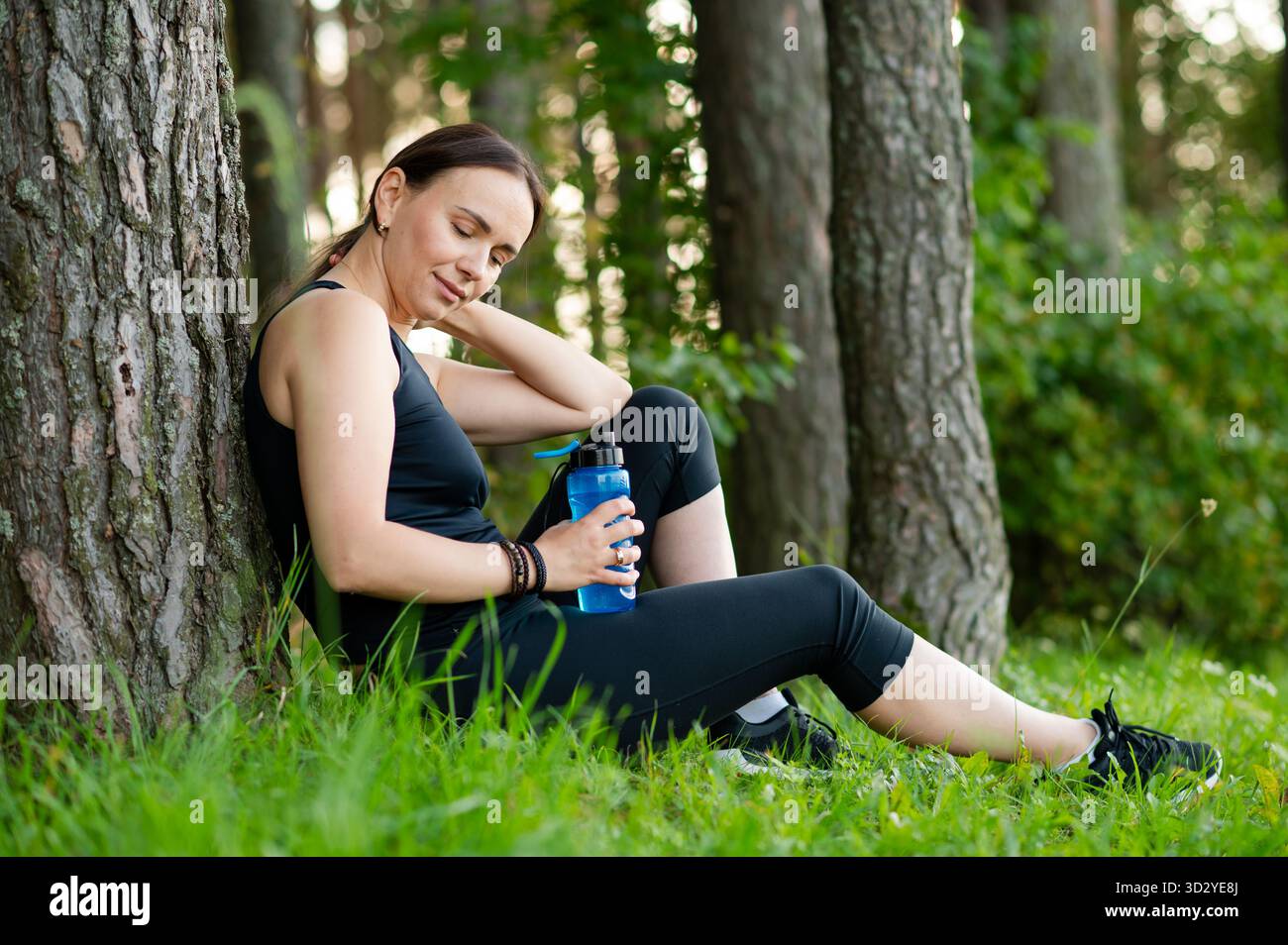 Belle jeune femme en vêtements de sport repose contre un arbre après avoir fait du jogging dans la forêt, tenant une bouteille d'eau et reprenant son souffle dans la nature paisible Banque D'Images