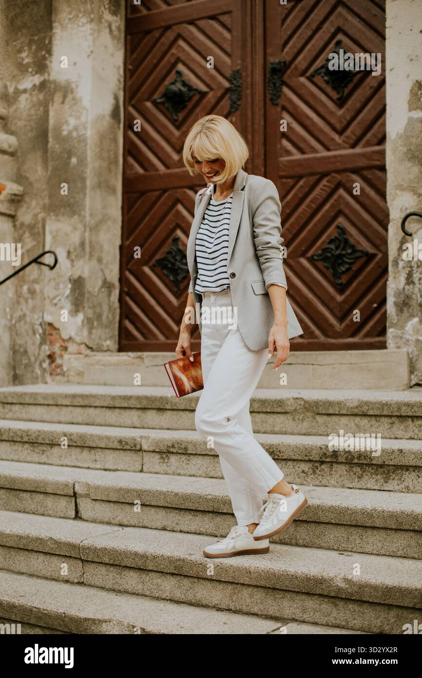 Une femme blonde portant un blazer, une chemise rayée et un pantalon blanc descend les marches de pierre. Elle porte une pochette près d'une porte en bois d'un bâtiment. Banque D'Images