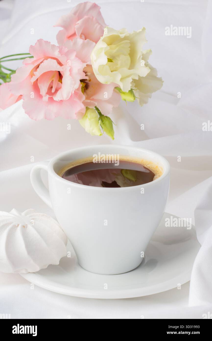 Une tasse à café blanche repose sur une soucoupe à côté d'une meringue molle. Les fleurs roses et blanches ajoutent une touche d'élégance sur un fond de tissu léger et fluide. Banque D'Images