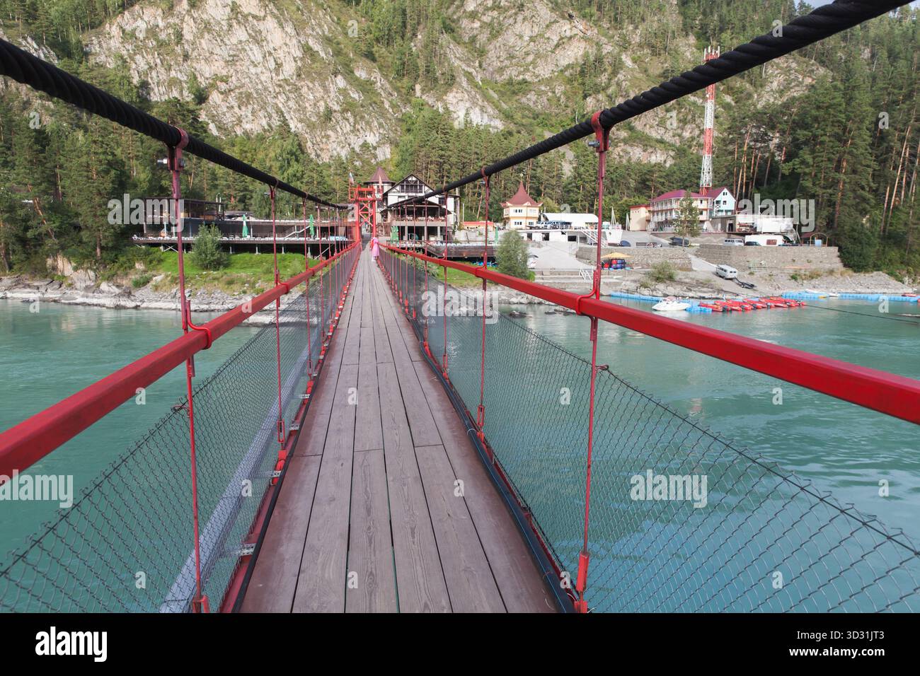 Un pont suspendu enjambe une rivière turquoise, guidant les voyageurs vers un village de montagne niché parmi les pins. Garde-corps rouges et filet de protection Banque D'Images