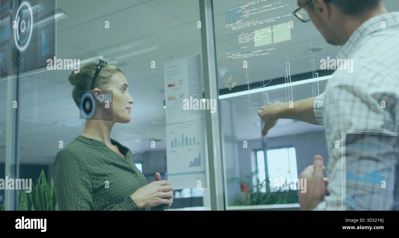 Deux collègues en tenue d'affaires pointant, regardant des graphiques et des superpositions sur la cloison de verre de bureau Banque D'Images