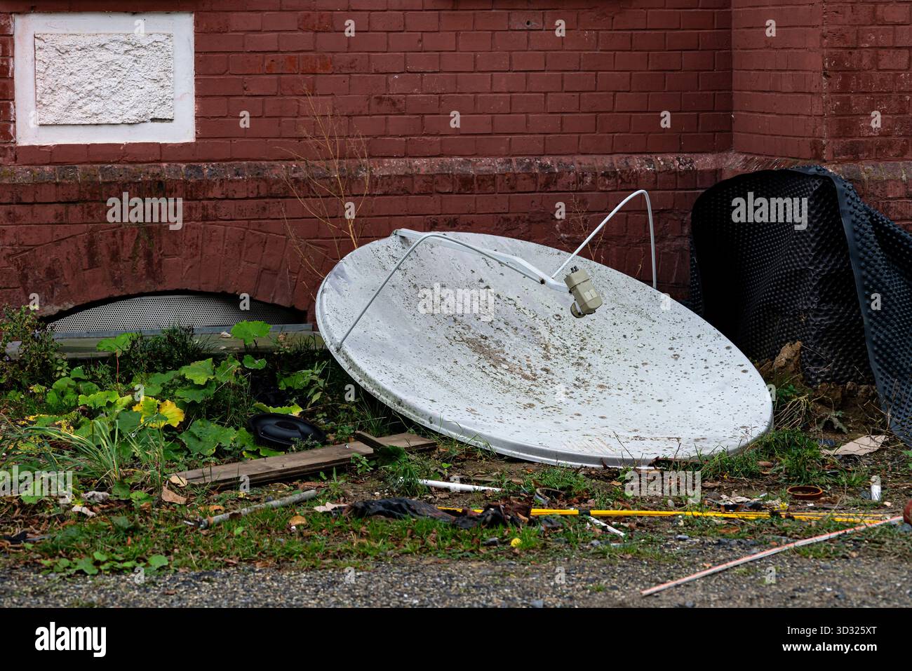 Une grande antenne parabolique cassée repose sur son côté à côté d'un bâtiment en briques. Autour, il est envahi par l'herbe et les débris, soulignant le St négligé Banque D'Images
