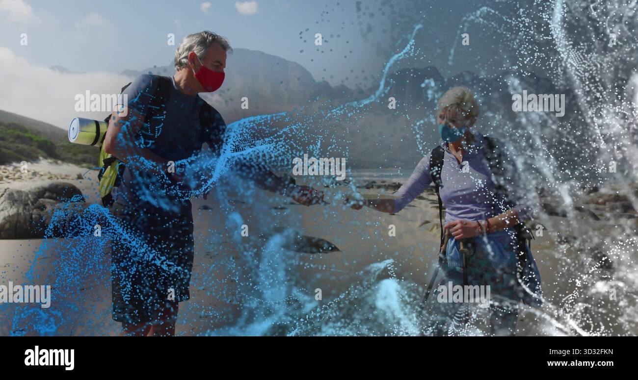 Randonneurs seniors debout portant des masques sur le bord de mer rocheux avec des bâtons de randonnée et une couche d'eau numérique Banque D'Images