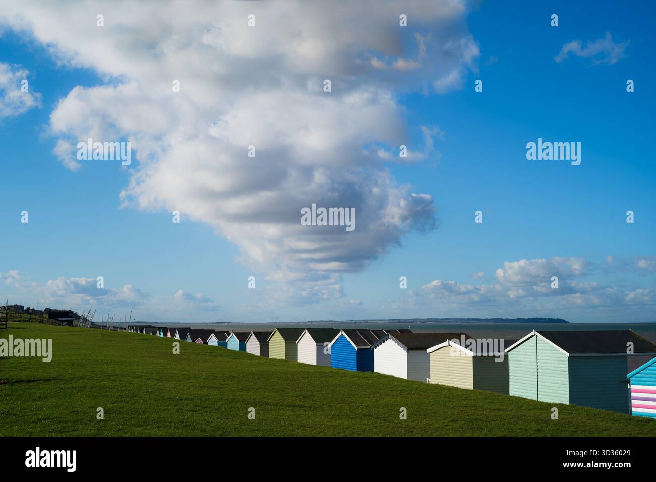Une rangée de cabanes de plage en bois colorées à Tankerton, Whitstable. Un grand nuage blanc domine le ciel bleu. L'île de Sheppey est à l'horizon. Banque D'Images