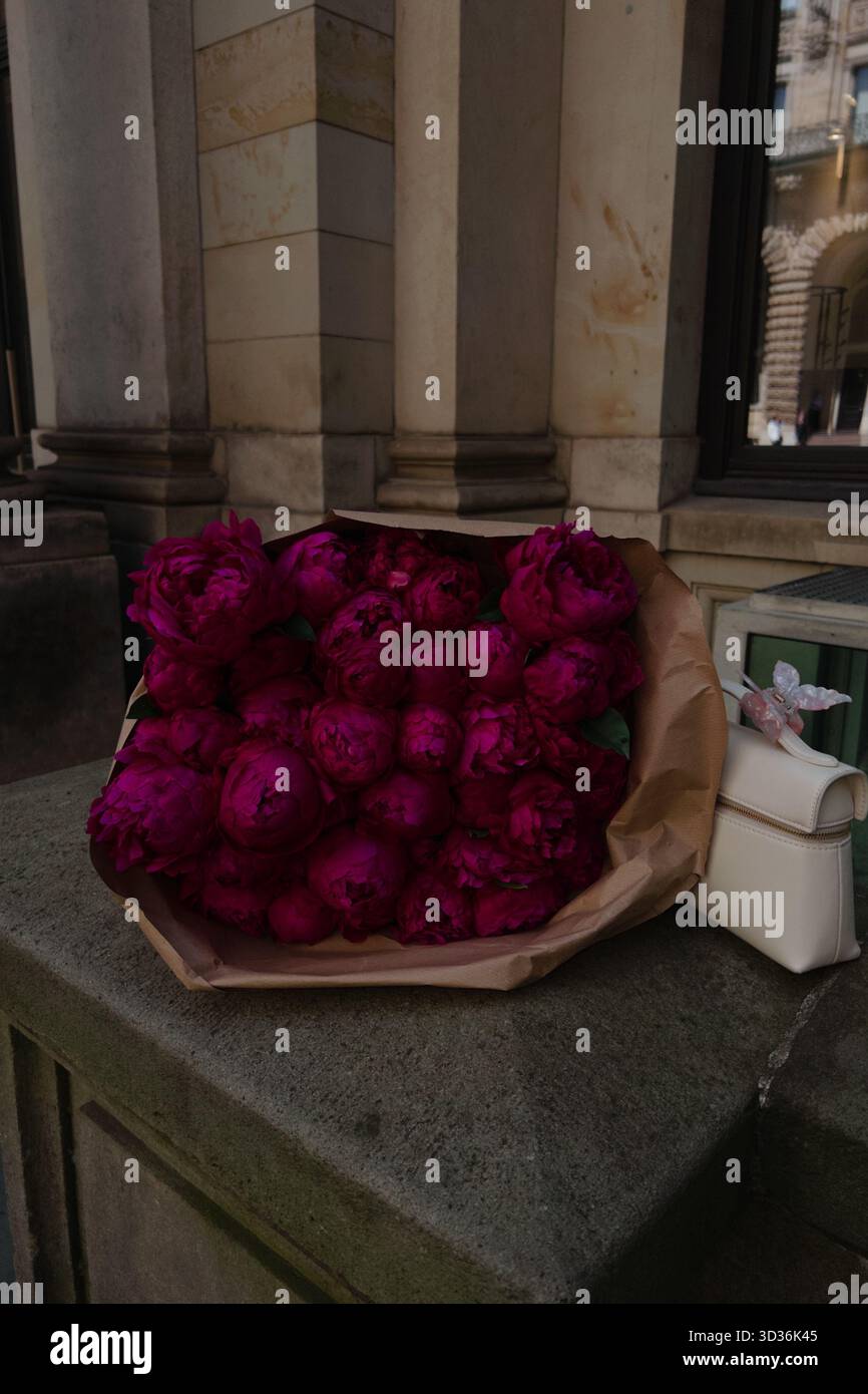 Un luxueux bouquet de pivoines magenta vibrantes enveloppées dans du papier brun, placé à côté d'un petit sac à main en cuir blanc sur un vieux rebord en pierre - Image de stock capturée avec un smartphone
