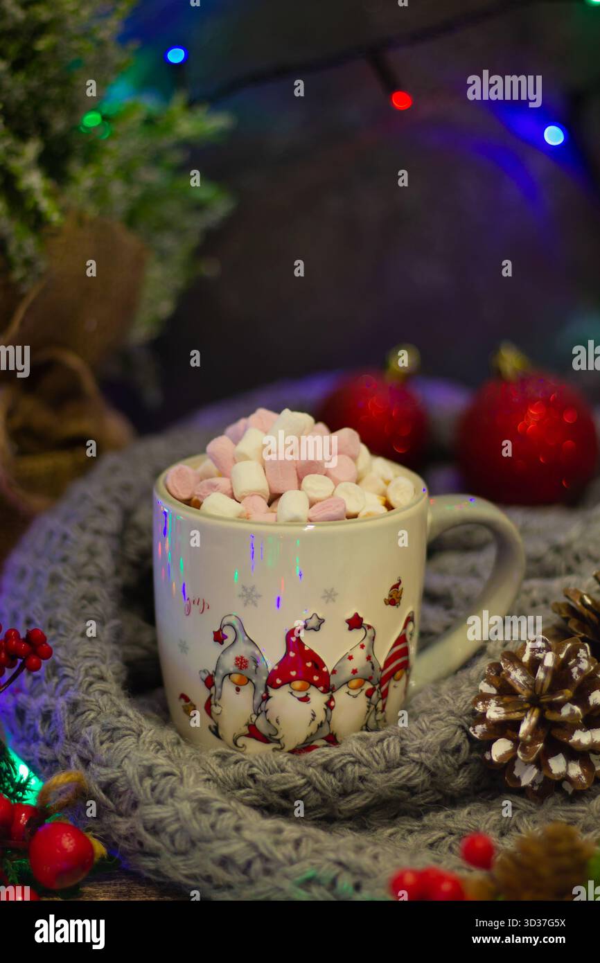 Une tasse confortable remplie de guimauves roses et blanches est entourée de pommes de pin, d'ornements festifs et de lumières scintillantes, capturant des vacances festives Banque D'Images