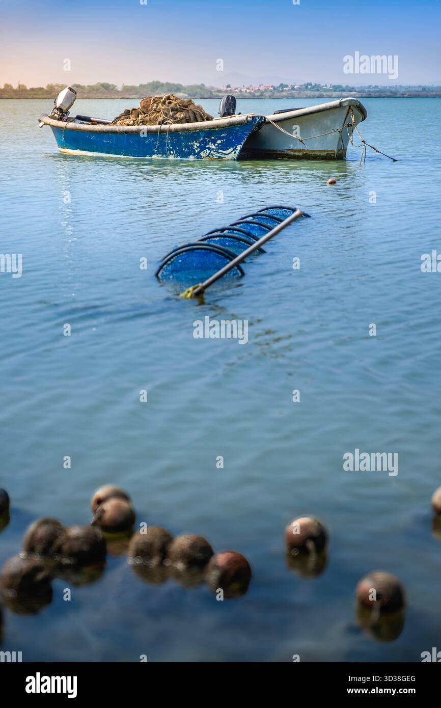Bateau de pêche bleu et blanc avec filets et moteur, ancré dans l'eau calme de la lagune, avec des bouchons flottants au premier plan, France Banque D'Images