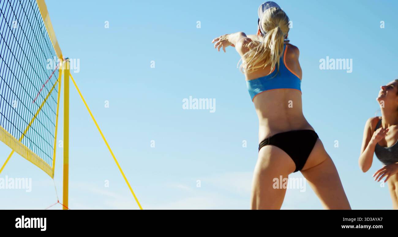 Athlète sautante frappant un volley bleu et blanc sur un terrain de plage, avec un filet jaune Banque D'Images