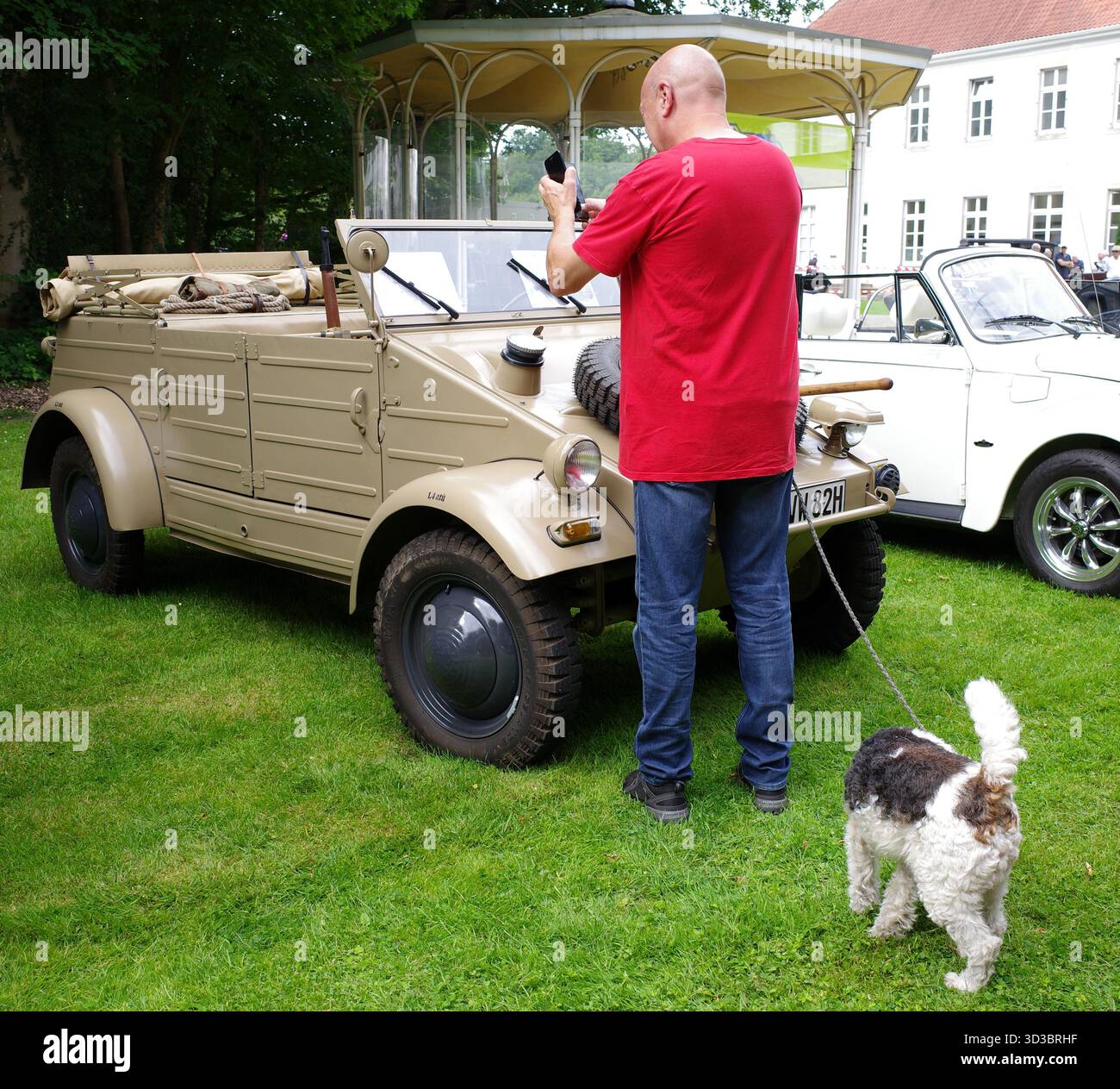 Bad Bentheim, Allemagne - 13 juillet 2025 Un homme avec un chien photographie une Volkswagen type 82 Kübelwagen avec son smartphone lors d'une rencontre de voitures anciennes Banque D'Images