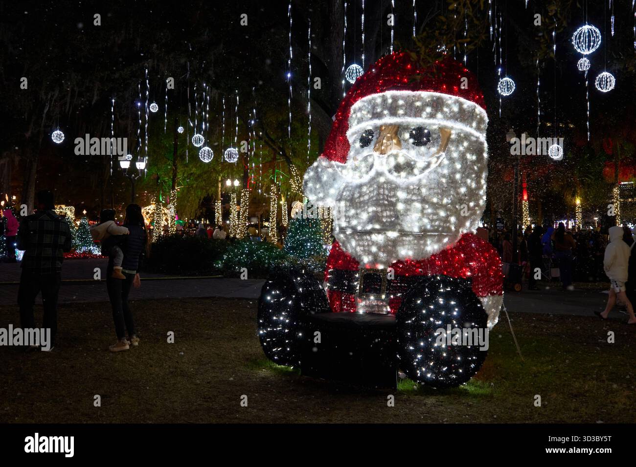 Père Noël géant fait de lumières LED brille dans un parc festif la nuit. Décoration de Noël avec neige, ornements et éclat lumineux autour des familles. Banque D'Images