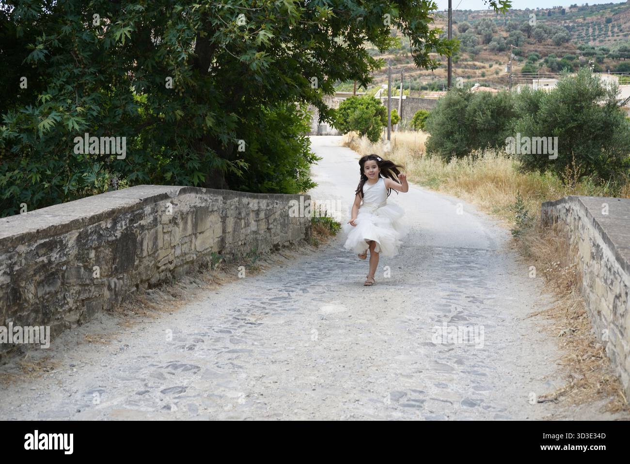 Une jeune fille joyeuse dans une robe blanche fluide court à travers le pont Apomarma en pierre capturant un moment d'été ludique dans la campagne grecque. Banque D'Images