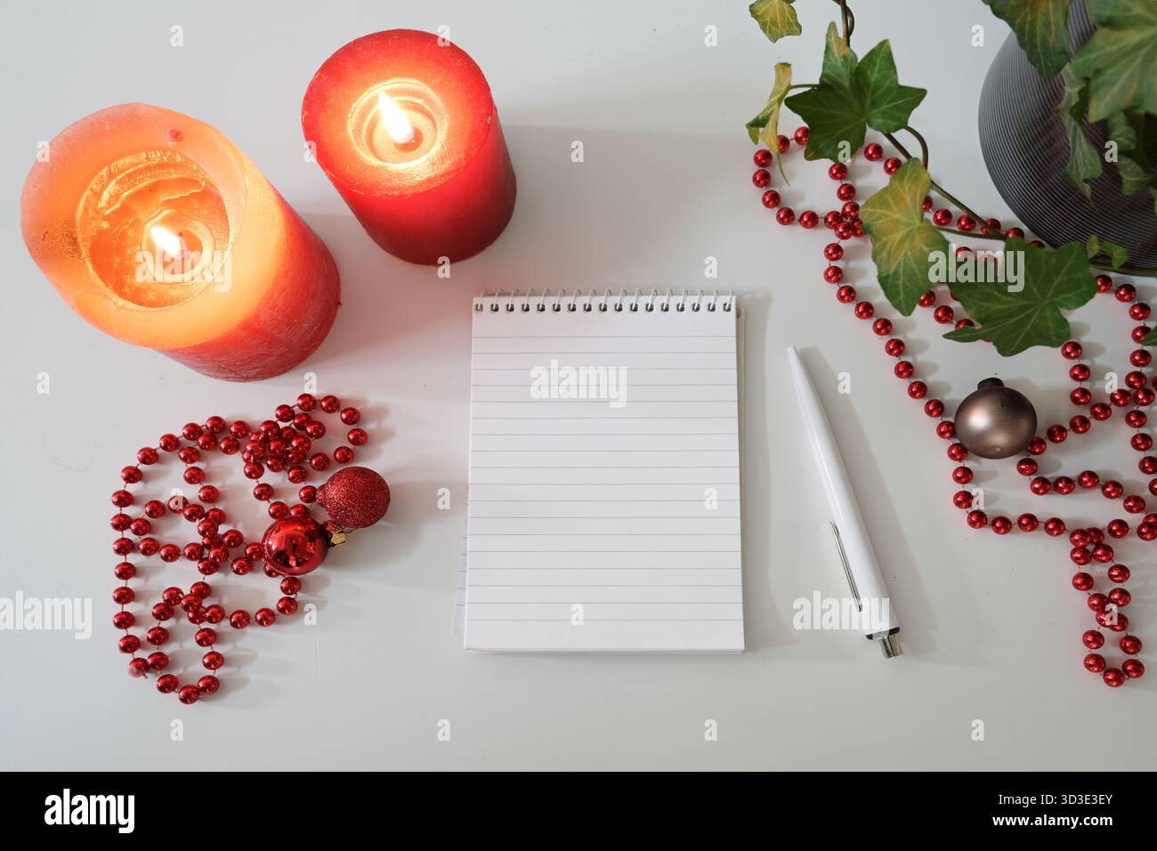Bloc-notes ligné, bougies rouges et décorations de Noël sur une table blanche, maquette pour une liste de choses à faire, liste de courses, ou liste de souhaits pour les vacances, copie s. Banque D'Images