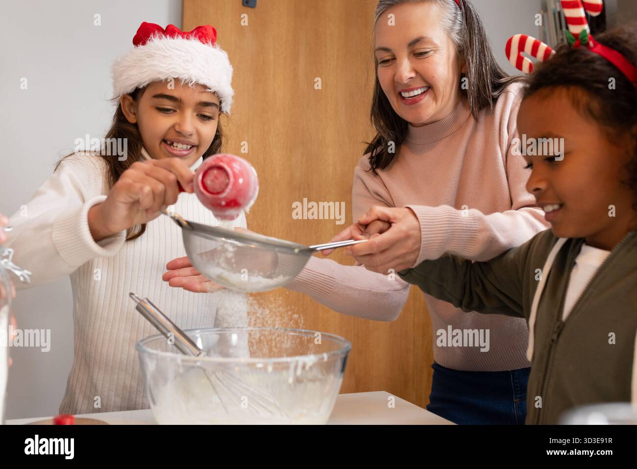 Famille diversifiée préparant le mélange de cuisson dans la cuisine à la maison en utilisant la tasse à mesurer rouge, le tamis et le bol Banque D'Images