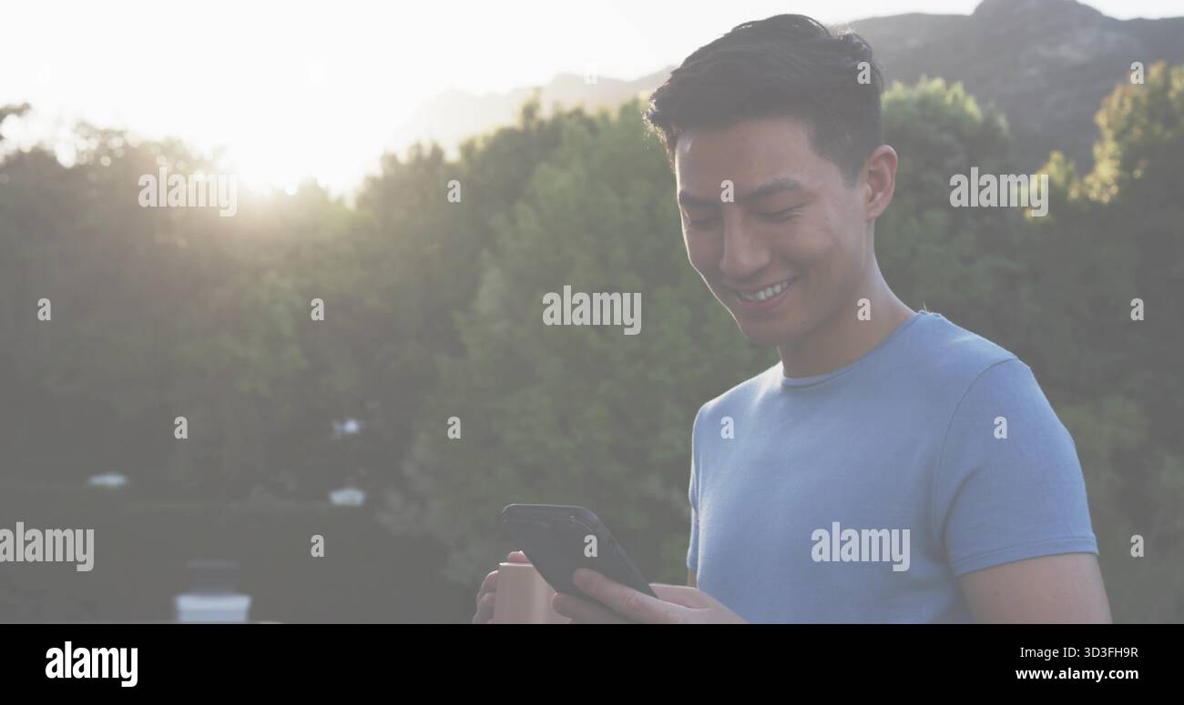 Homme asiatique souriant en t-shirt bleu clair tenant smartphone sur terrasse, avec tasse à café, espace copie Banque D'Images