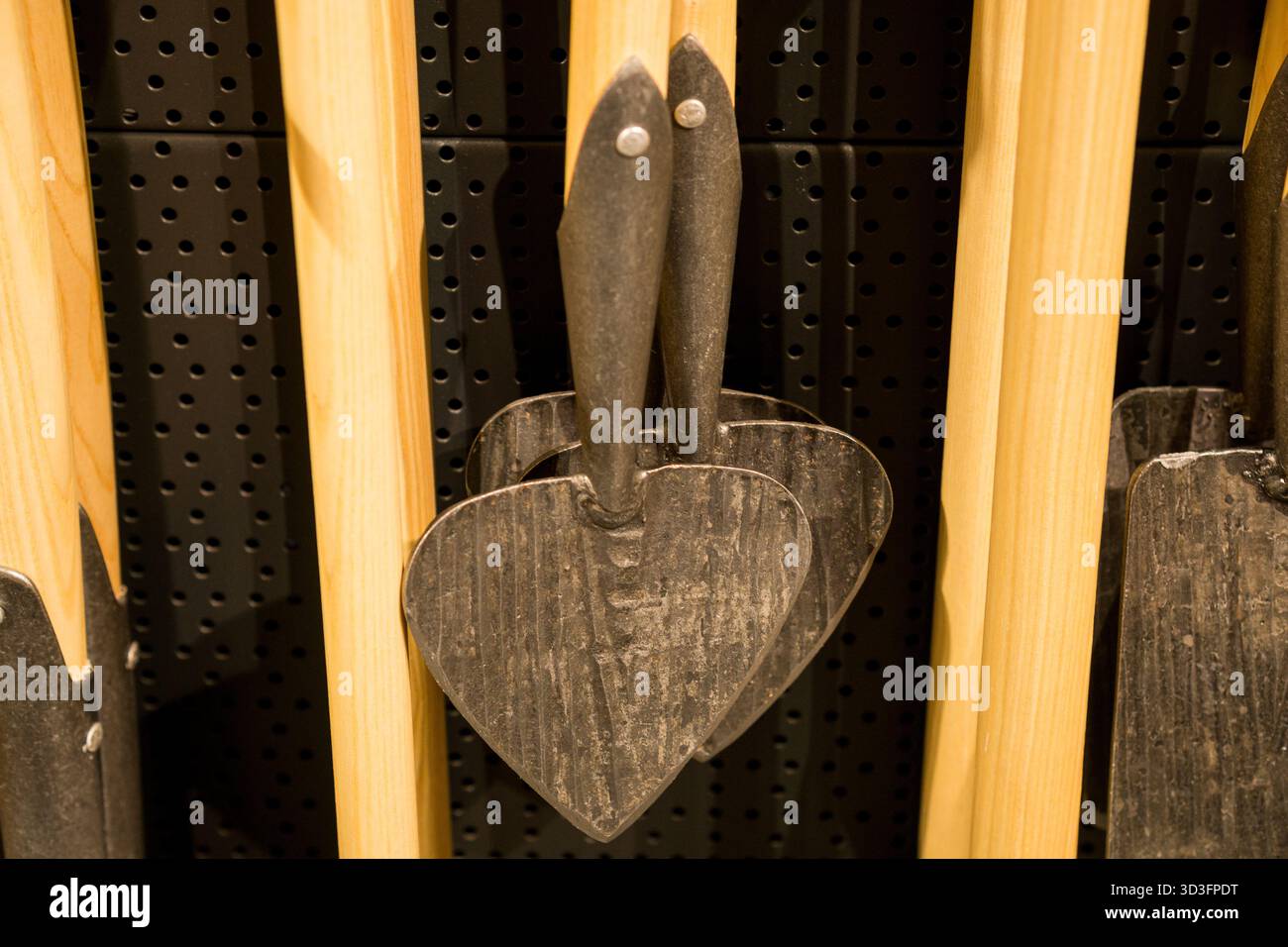 Gros plan de pelles de jardin métalliques en forme de cœur suspendues à des poignées en bois sur fond perforé noir dans une quincaillerie. Banque D'Images