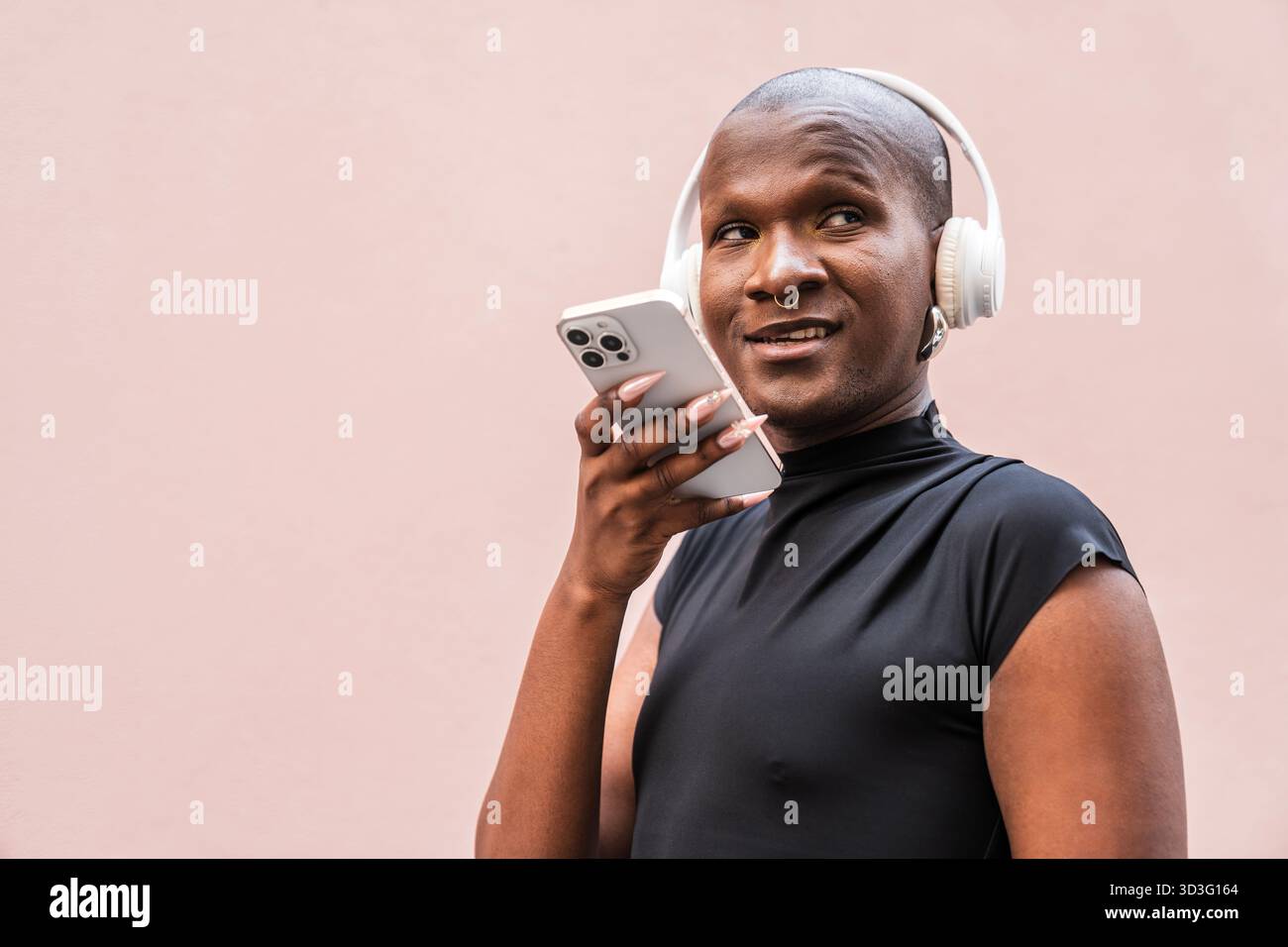 Un jeune homme LGBTQ se tient confiant, souriant et interagissant avec son smartphone tout en portant des écouteurs sur un fond neutre, embrassant con Banque D'Images