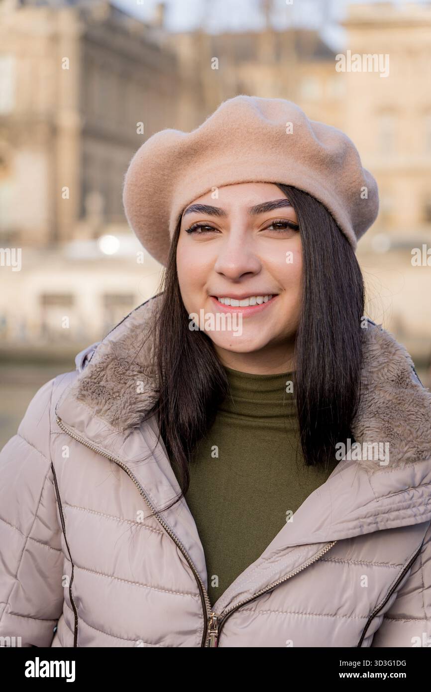 Femme élégante à Paris portant un béret beige confortable et un manteau d'hiver, souriant chaleureusement à l'extérieur avec un bâtiment parisien classique floue en arrière-plan. Banque D'Images