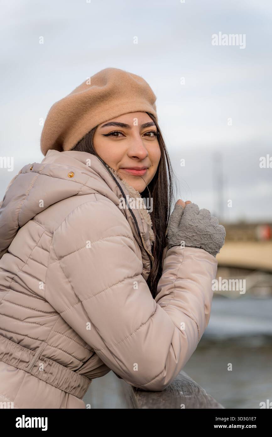 Une femme vêtue d'un béret beige et d'une veste bouffante s'appuie sur une balustrade, profitant d'une journée d'hiver vigoureuse au bord de la Seine à Paris, mettant en valeur le style français classique Banque D'Images