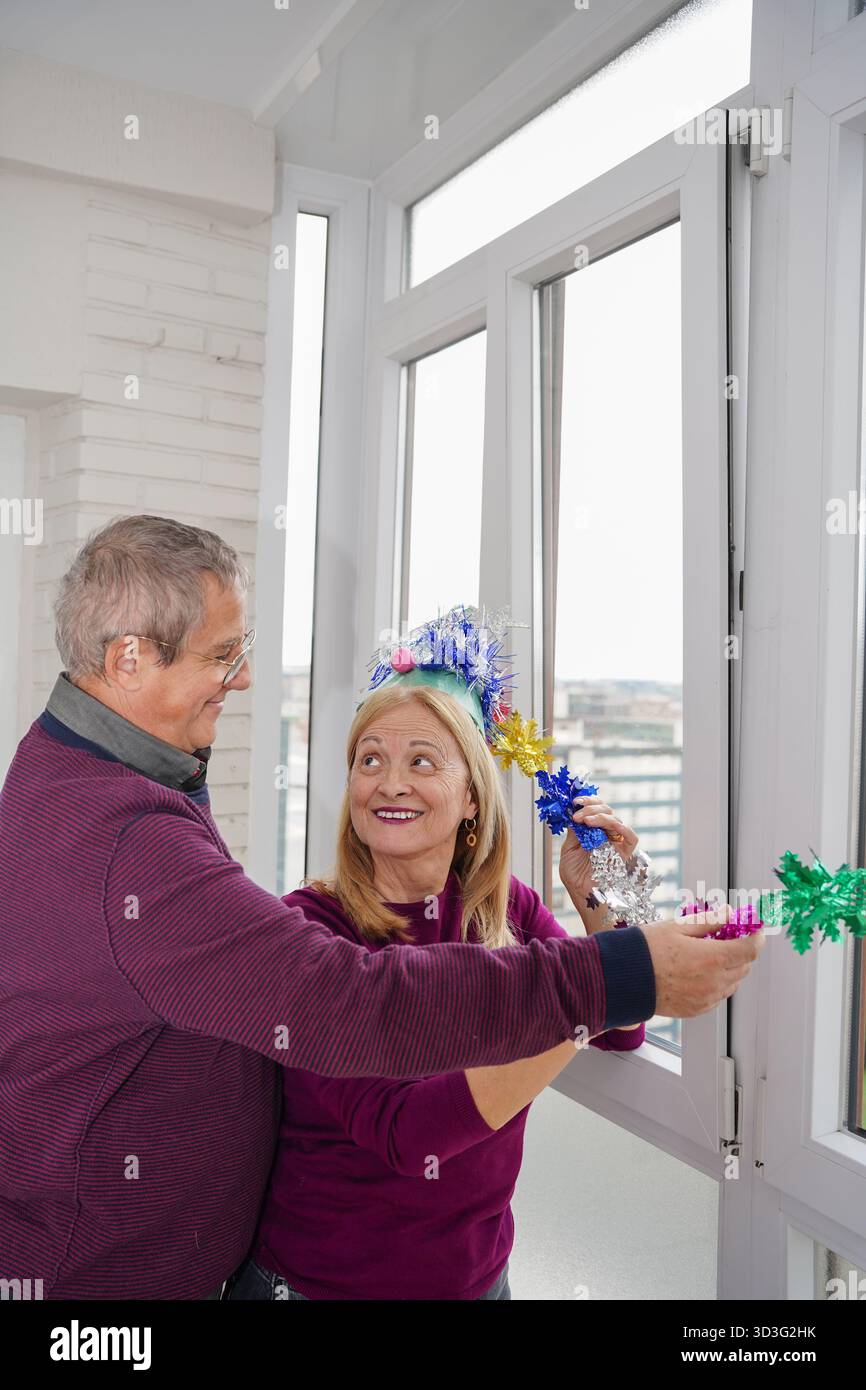 Un couple âgé joyeux s'engageant dans un esprit festif, décorant leur maison avec des guirlandes colorées, incarnant la chaleur des traditions de Noël et de famil Banque D'Images