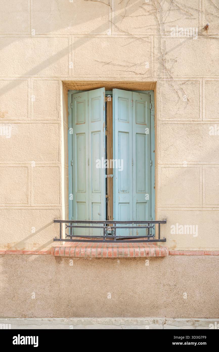 Fenêtre simple avec volets en bois bleu clair légèrement entrouverts, dans un mur neutre en stuc avec un rebord en briques rouges et une balustrade en fer forgé Banque D'Images