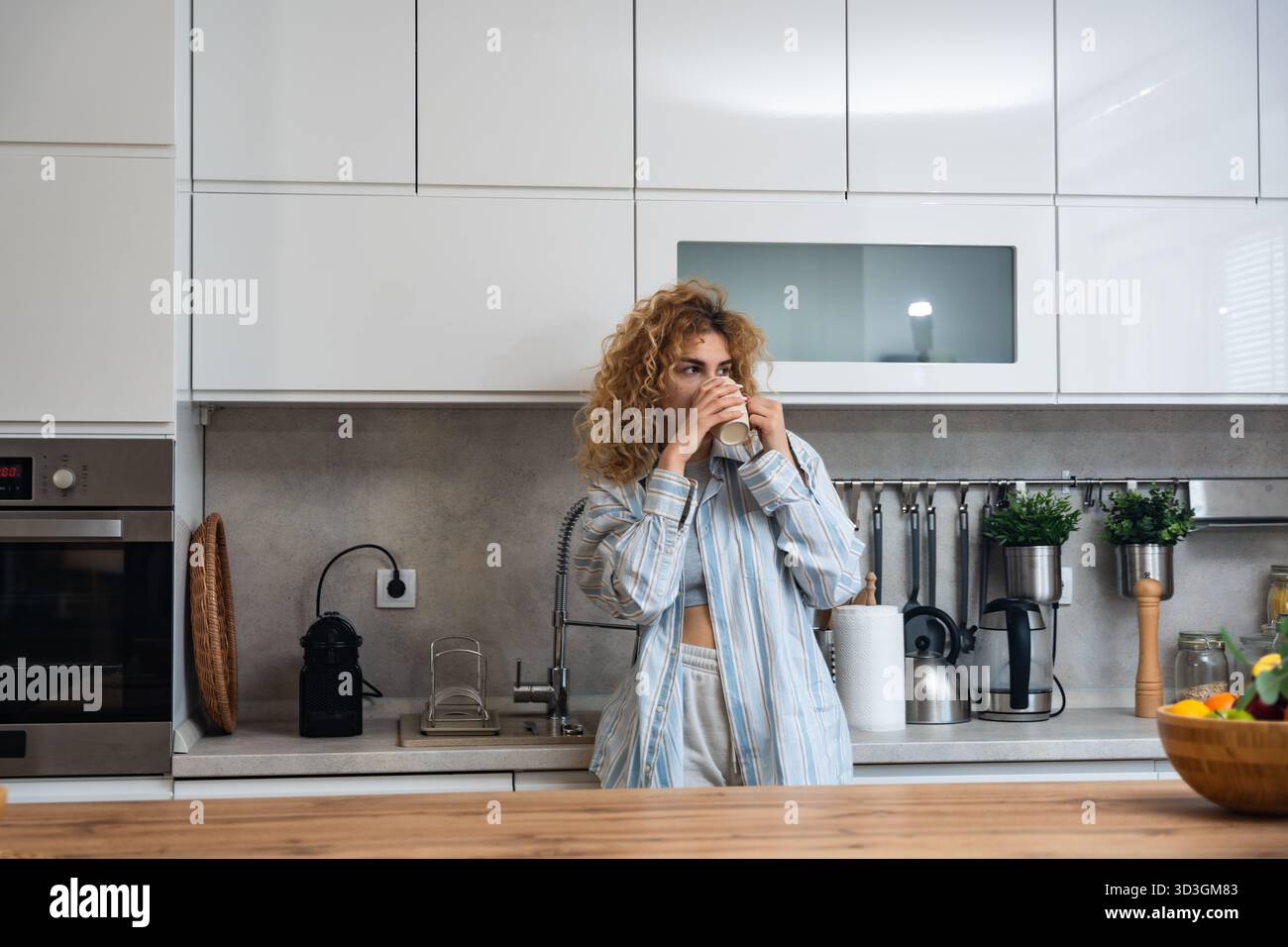 Jeune femme endormie dans la cuisine à la maison sirotant du café lente routine matinale dans des vêtements de détente décontractés, chemise rayée et boucles désordonnées, profitant d'une pause tranquille par Banque D'Images