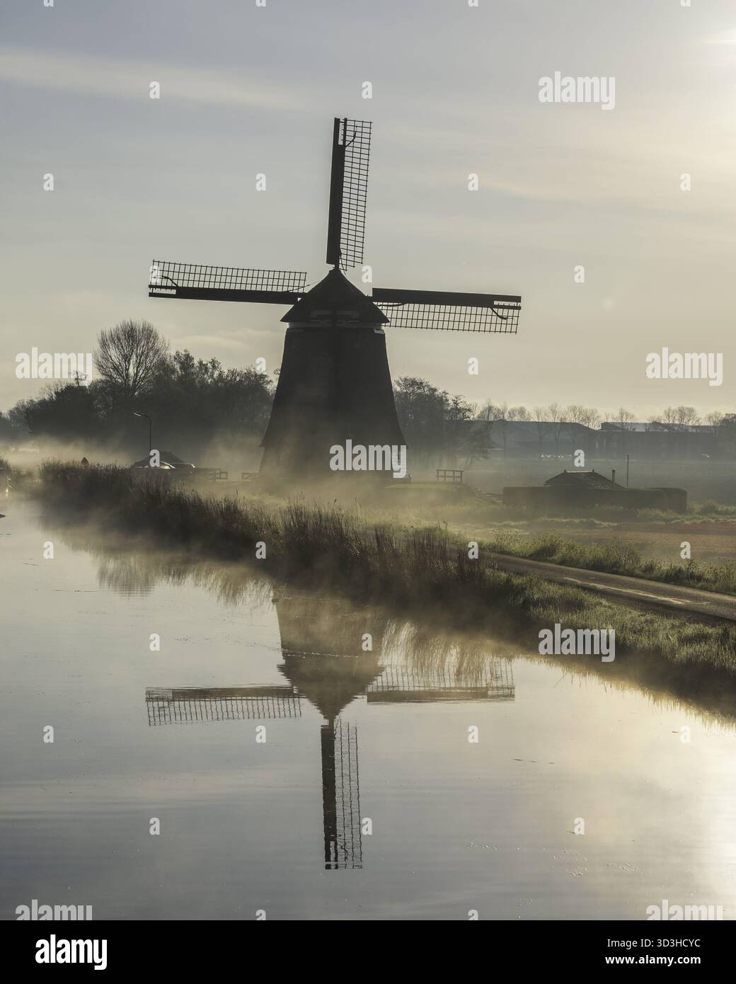 Vue d'un moulin à vent tranquille reflété dans les eaux calmes d'un canal brumeux à l'aube, peignant un paysage serein, Obdam, Hollande du Nord, pays-Bas. Banque D'Images