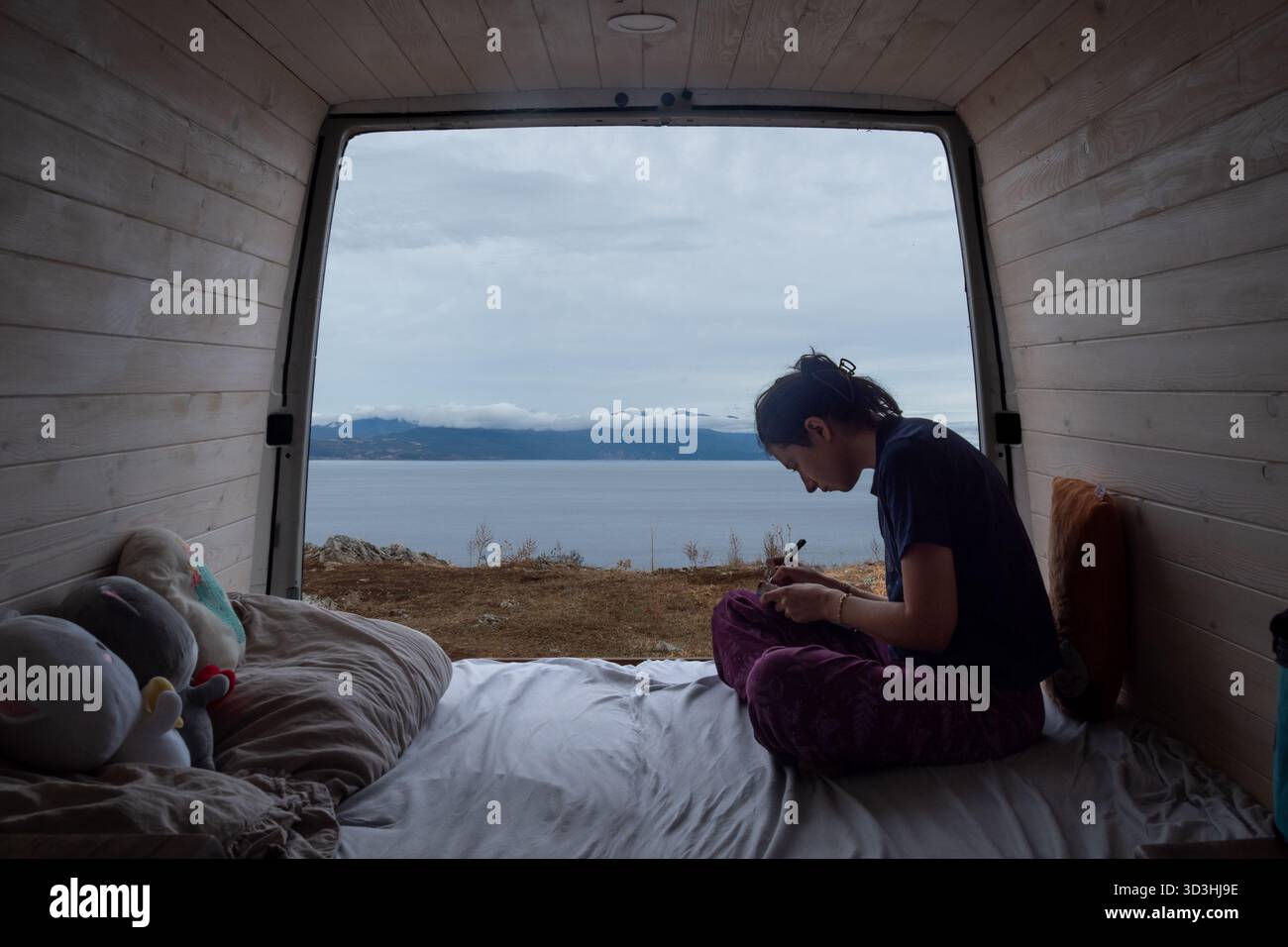 Jeune femme assise dans un camping-car, écrivant ou dessinant tout en regardant un paysage de lac calme. Banque D'Images